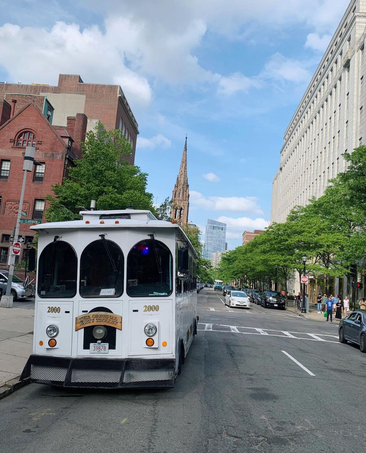 White sightseeing trolley parked on a leafy downtown city street with a tall historic church spire and modern office buildings under a bright blue sky.