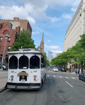 White sightseeing trolley parked on a leafy downtown city street with a tall historic church spire and modern office buildings under a bright blue sky.