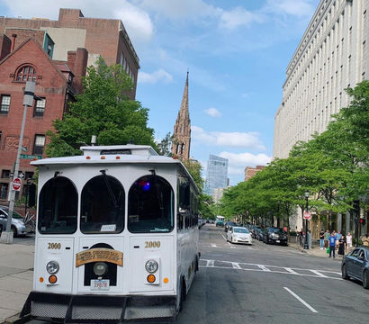 White sightseeing trolley parked on a leafy downtown city street with a tall historic church spire and modern office buildings under a bright blue sky.