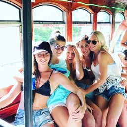Five smiling friends in swimsuits and denim shorts posing together inside a vintage wooden trolley with arched windows on a sunny summer ride.