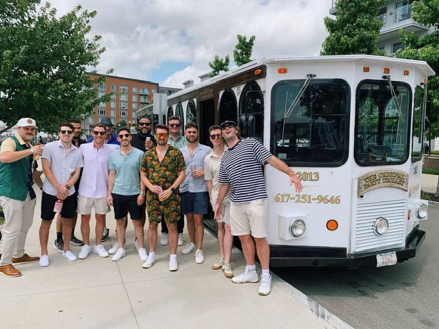 Group of men in casual summer outfits smiling and posing beside a white vintage-style trolley on a tree-lined city sidewalk with apartment buildings and a partly cloudy sky in the background.