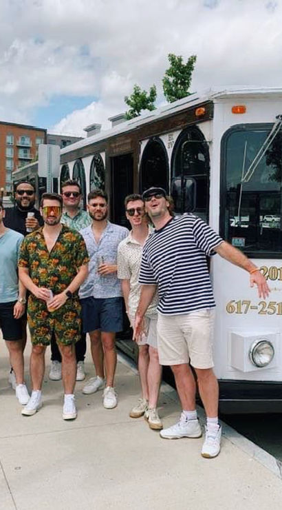 Group of men in casual summer outfits smiling and posing beside a white vintage-style trolley on a tree-lined city sidewalk with apartment buildings and a partly cloudy sky in the background.