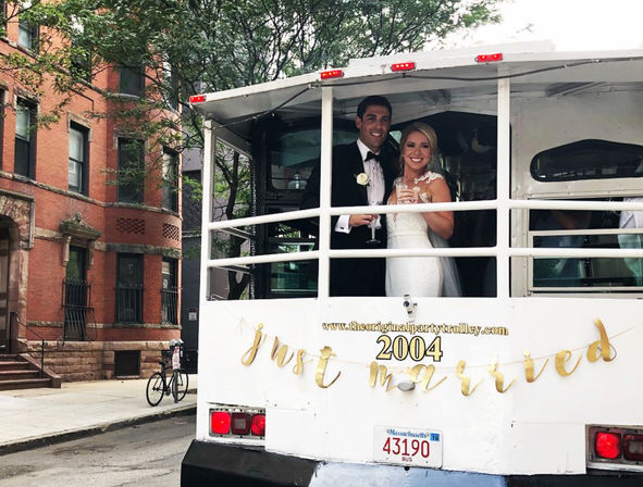 Bride and groom in formal wedding attire smiling and toasting aboard a white party trolley decorated with a gold "just married" banner on a city brownstone-lined street.