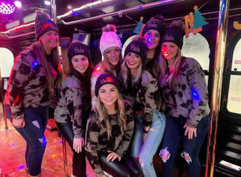 Bachelorette party on a party bus — a group of friends in matching camo sweatshirts and knit beanies (most read "Babe", one reads "Bride") posing under colorful disco lights and poles.