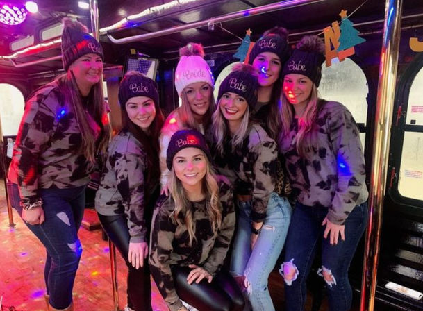 Bachelorette party on a party bus — a group of friends in matching camo sweatshirts and knit beanies (most read "Babe", one reads "Bride") posing under colorful disco lights and poles.
