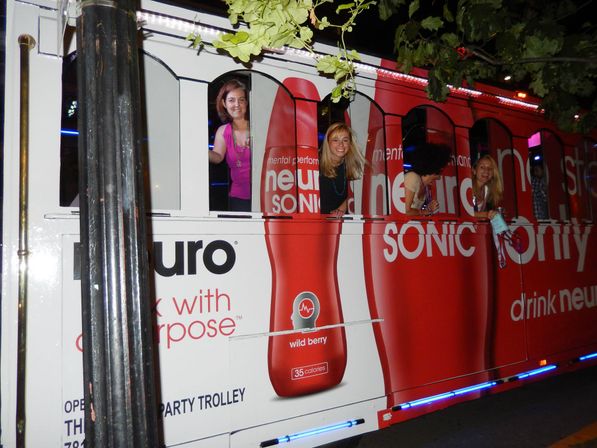 Three smiling women leaning from windows of a red nighttime party trolley wrapped in large bottled beverage graphics advertising a wild berry drink, lit by streetlights and blue LED trim on an urban street.