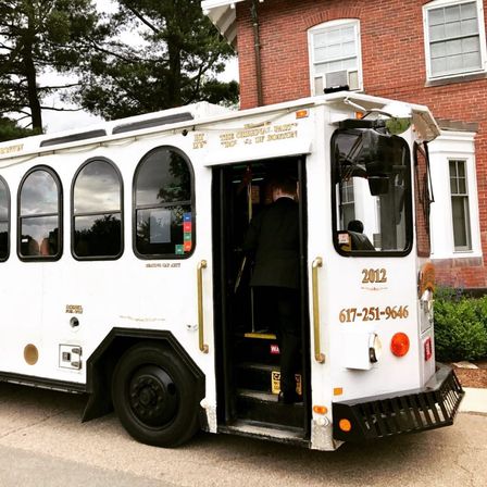 White vintage-style Boston trolley with arched windows and brass handrails parked by a red brick building, a passenger in a suit boarding through the open door