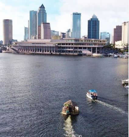 Two small boats, one with a tiki-hut deck, crossing Tampa waterfront toward a downtown skyline with a waterfront convention center and high-rise buildings on a sunny day