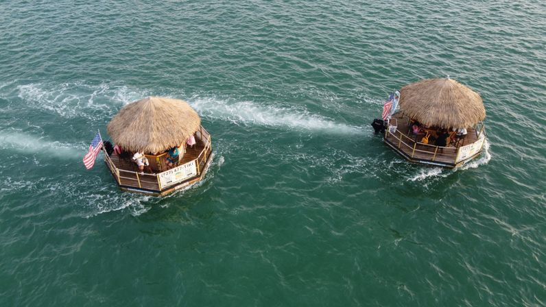 Aerial view of two round floating tiki-hut boats with thatched roofs and American flags, passengers enjoying a tiki-raft boat tour leaving wake trails across teal coastal water.