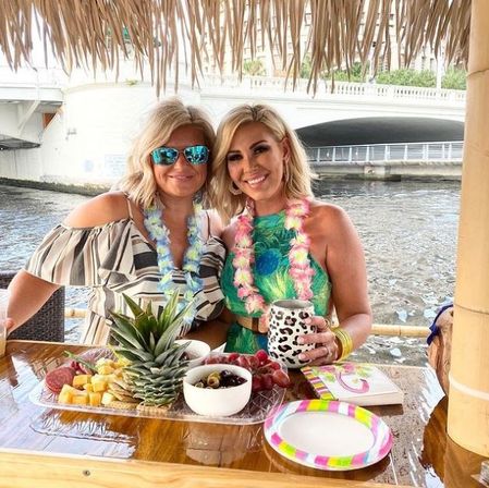 Two smiling women in leis at a waterfront tiki hut enjoying a tropical fruit platter with pineapple centerpiece, cheese, olives and a patterned drink cup by a river bridge