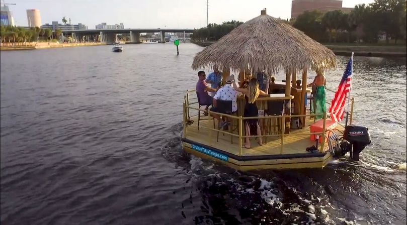Group of people enjoying a thatched-roof tiki bar boat on an urban river, American flag fluttering, bridge and waterfront buildings in the background.