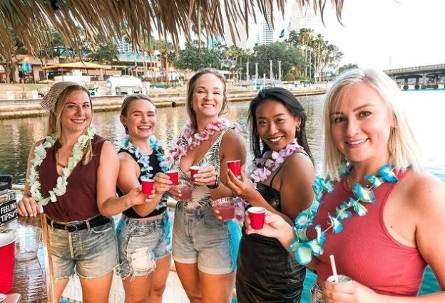 Five women wearing colorful leis smiling and toasting with cocktails and red cups at a waterfront tiki-style bar by a marina with palm trees and boats in the background.