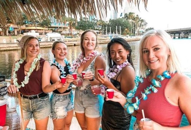 Five women wearing colorful leis smiling and toasting with cocktails and red cups at a waterfront tiki-style bar by a marina with palm trees and boats in the background.