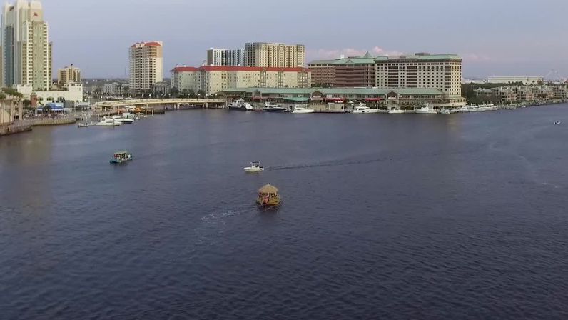 Aerial view of a riverfront skyline and marina with hotels and yachts, small colorful tour boats and other vessels cruising across calm water.