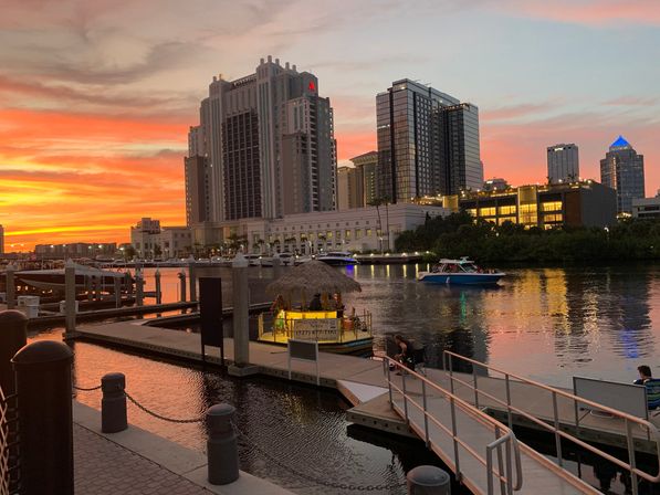 Sunset over a waterfront city skyline with glowing orange skies, high-rise buildings, boats at a marina, a lit dockside tiki hut and reflections on calm water.