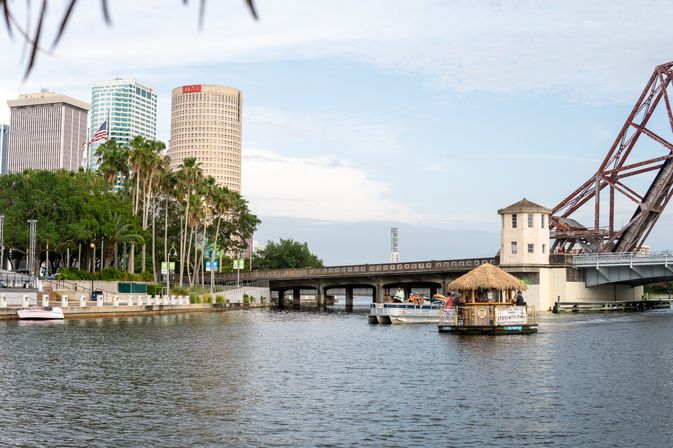 Tampa waterfront on the Hillsborough River with palm trees, a cylindrical downtown skyscraper, a rusty steel drawbridge, and boats including a thatched-roof tiki bar floating by a low concrete bridge.