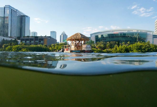 Low-angle half-submerged shot of a floating tiki-hut bar with people under a thatch roof, calm water, green shoreline and a downtown waterfront skyline of glass high-rises and an arena on a sunny day.