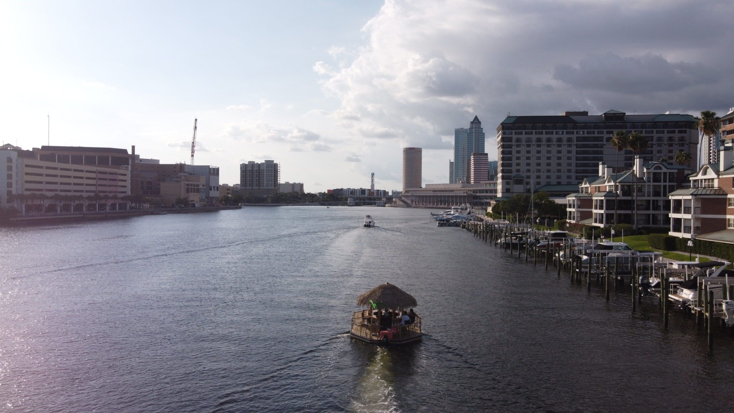 Tiki-style thatched-roof pontoon boat cruising down a downtown waterfront channel toward a skyline of high-rise buildings and marina docks under a partly cloudy sky