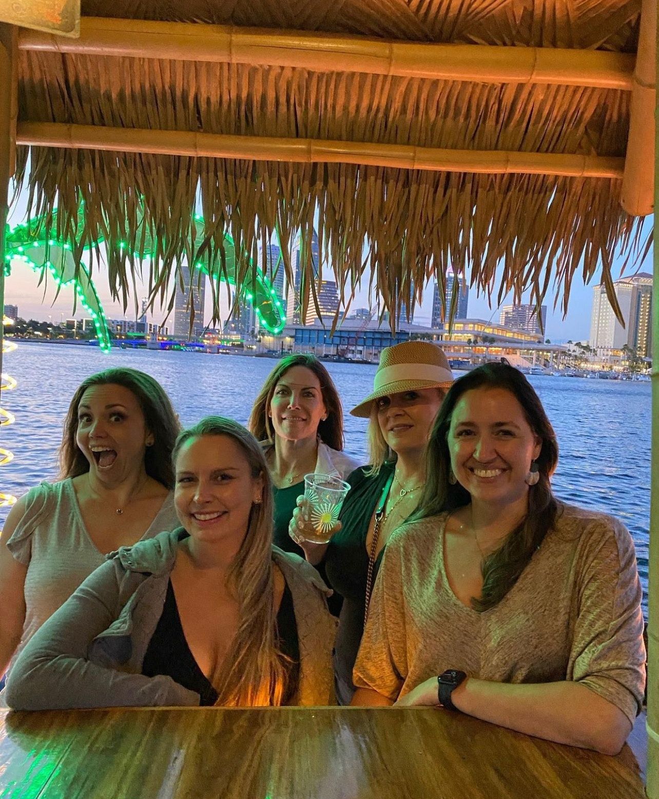 Group of five women smiling under a thatched tiki hut at a waterfront bar, neon-green lights and an urban skyline at dusk reflected on the harbor, wooden table in the foreground