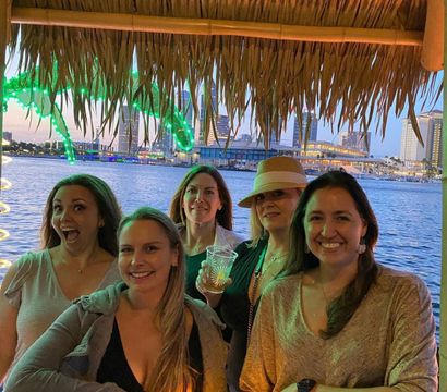 Group of five women smiling under a thatched tiki hut at a waterfront bar, neon-green lights and an urban skyline at dusk reflected on the harbor, wooden table in the foreground