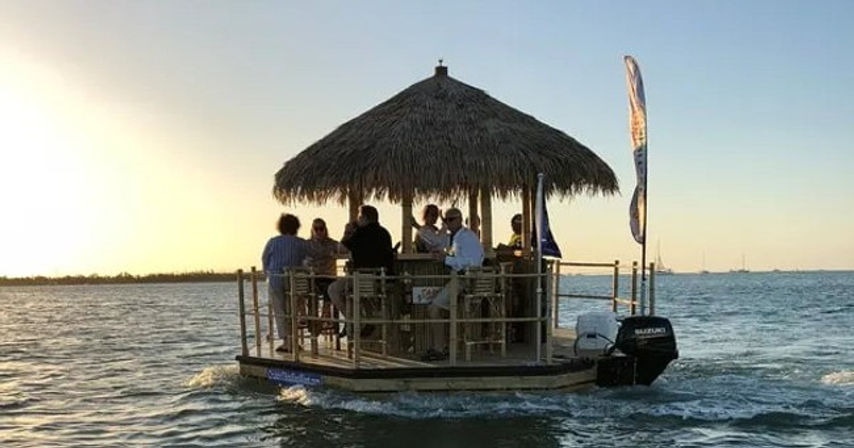 Motorized floating tiki bar with thatched roof and flag carrying a group of people enjoying drinks at sunset on calm coastal waters