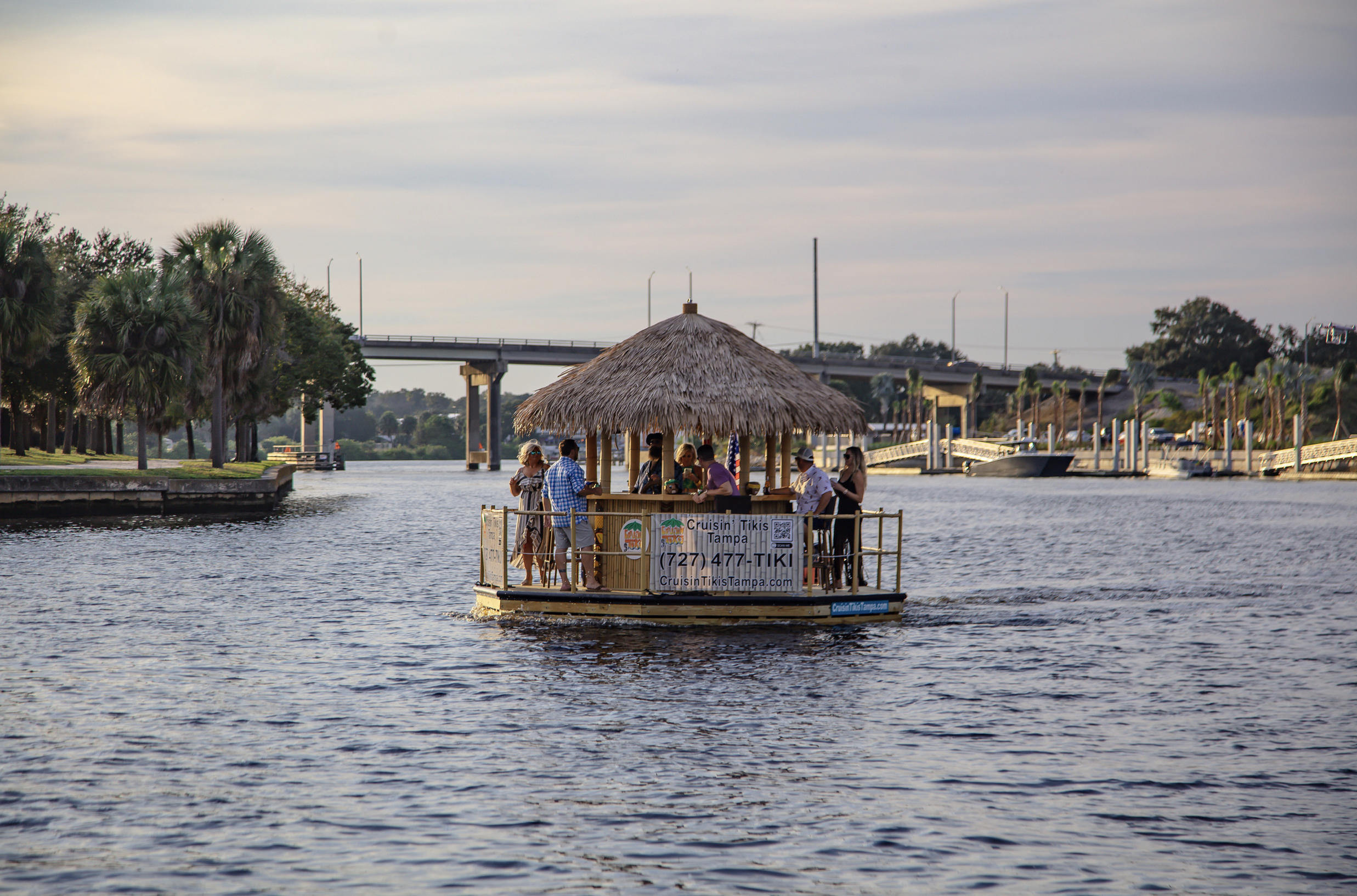Thatched-roof tiki boat with a group of people cruising a calm Tampa waterfront at golden hour, palm trees and a bridge in the background.
