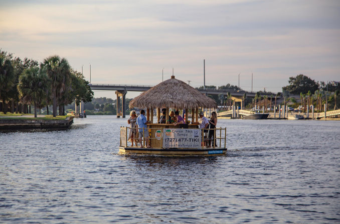 Thatched-roof tiki boat with a group of people cruising a calm Tampa waterfront at golden hour, palm trees and a bridge in the background.