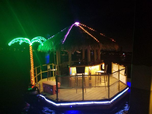 Neon-lit floating tiki bar with a thatched roof, LED palm tree and string lights glowing on a small deck over dark water at night