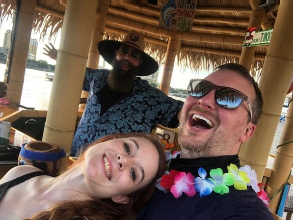 Three people laughing for a selfie inside a thatched-roof tiki bar on the waterfront, wearing colorful leis, sunglasses, and a wide-brim hat with a city skyline across the water.