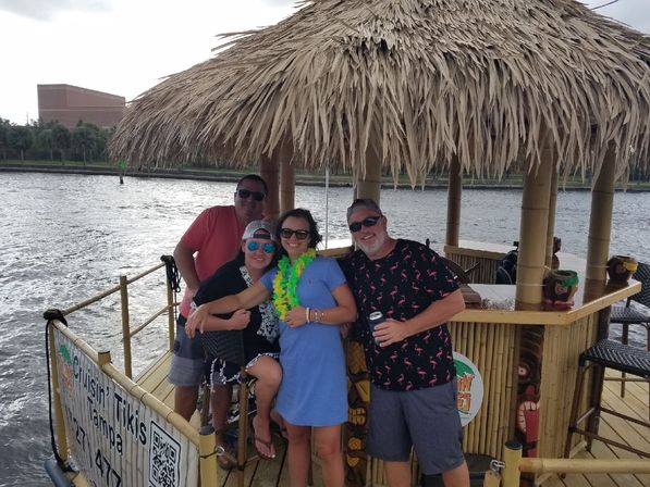Group of four adults wearing leis and sunglasses smiling aboard a bamboo tiki-bar boat with a thatched roof on the Tampa riverfront