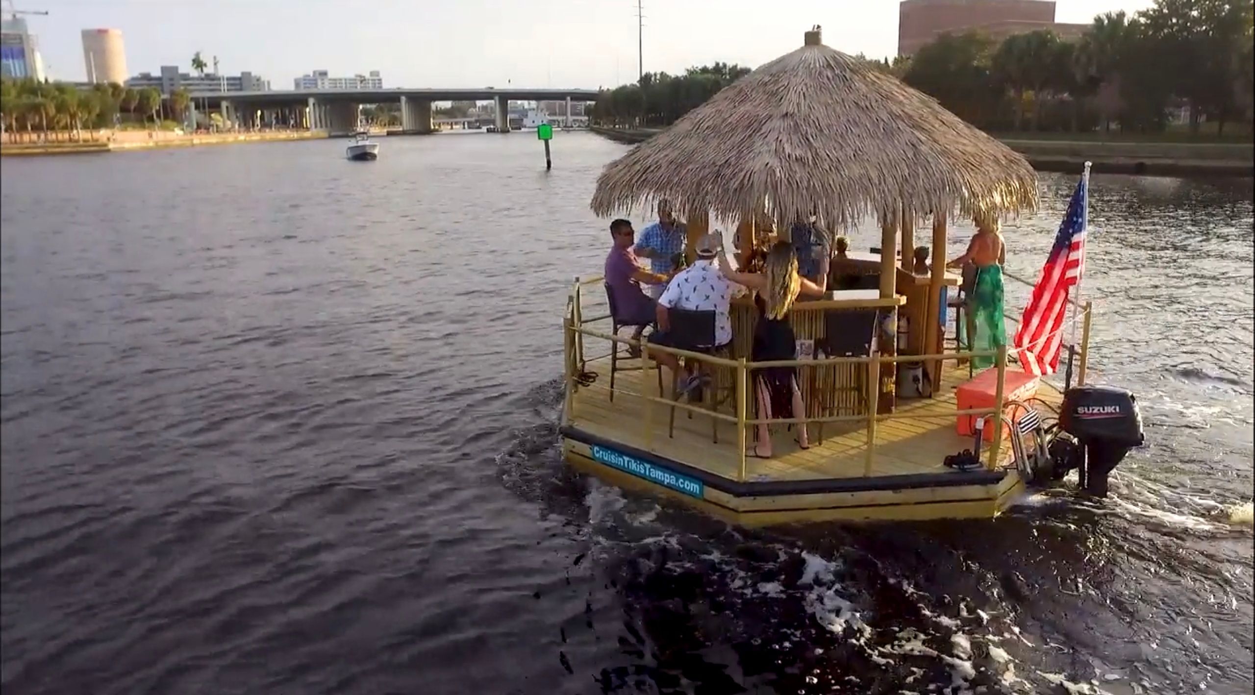Group enjoying a tiki-style party boat with a thatched roof and American flag cruising a city river past a bridge and waterfront buildings