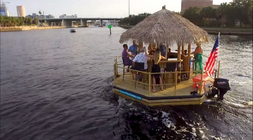 Group enjoying a tiki-style party boat with a thatched roof and American flag cruising a city river past a bridge and waterfront buildings