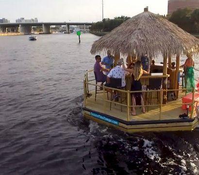 Group enjoying a tiki-style party boat with a thatched roof and American flag cruising a city river past a bridge and waterfront buildings