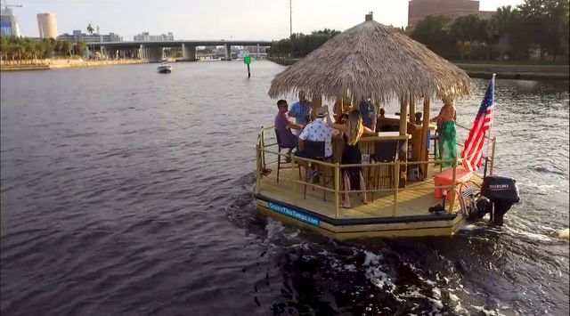 Group enjoying a tiki-style party boat with a thatched roof and American flag cruising a city river past a bridge and waterfront buildings