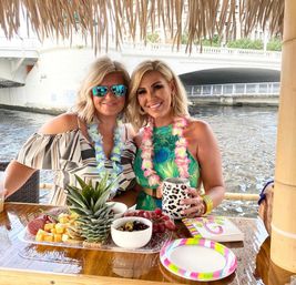 Two smiling women wearing floral leis at a waterfront tiki bar under a thatched roof, enjoying tropical drinks and a charcuterie platter with pineapple, cheese, grapes and olives by a river bridge.