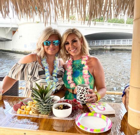 Two smiling women wearing floral leis at a waterfront tiki bar under a thatched roof, enjoying tropical drinks and a charcuterie platter with pineapple, cheese, grapes and olives by a river bridge.