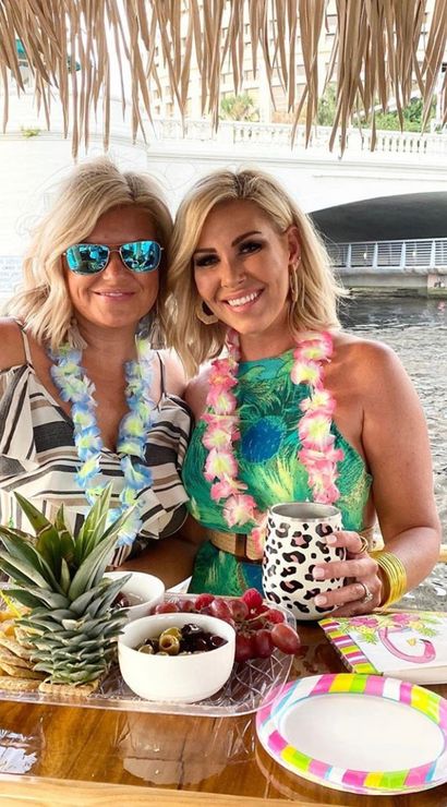 Two smiling women wearing floral leis at a waterfront tiki bar under a thatched roof, enjoying tropical drinks and a charcuterie platter with pineapple, cheese, grapes and olives by a river bridge.