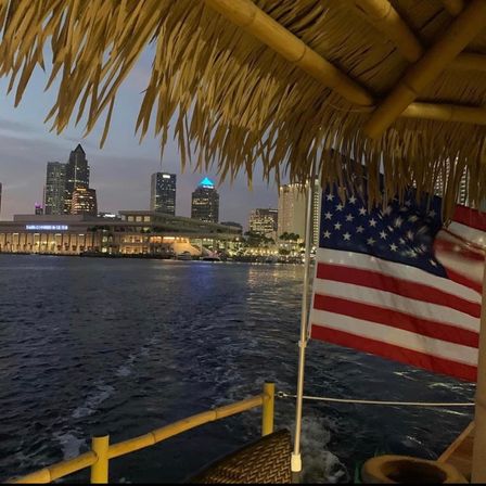 Tiki‑roofed boat view of a waterfront city skyline at dusk with an American flag fluttering over rippling harbor water and glowing building lights.