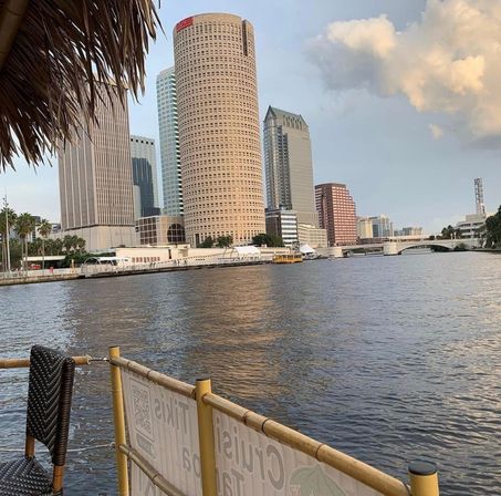 Tiki-style waterside patio view of downtown Tampa skyline with a round high-rise and bridge reflecting on the Hillsborough River at golden hour
