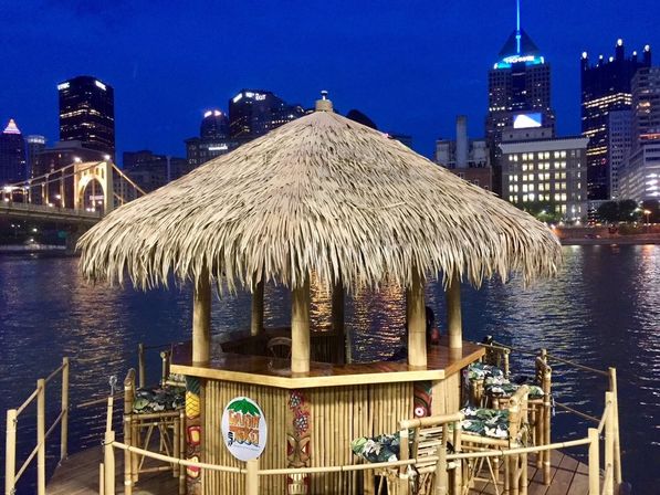 Thatched-roof tiki bar on a floating bamboo dock with cushioned stools, river reflections and an illuminated city skyline and arched bridge at dusk.