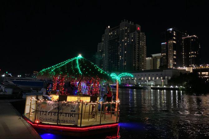 Colorful tiki boat decked in green and multicolored lights at night on the Tampa waterfront, reflecting on the water with illuminated downtown skyline in the background.