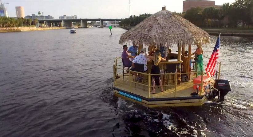 People enjoying a tiki-style pontoon boat with a thatched roof and American flag cruising an urban riverfront lined with palm trees past a bridge and city skyline.