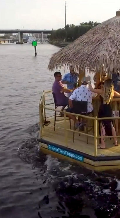 People enjoying a tiki-style pontoon boat with a thatched roof and American flag cruising an urban riverfront lined with palm trees past a bridge and city skyline.