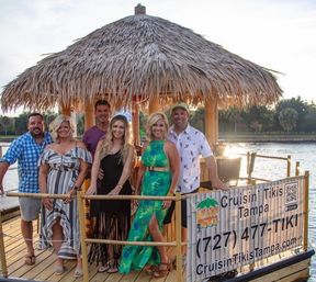 Six adults posing on a thatched-roof tiki-style boat with bamboo railings docked on a Tampa waterfront at sunset, smiling in colorful summer outfits.