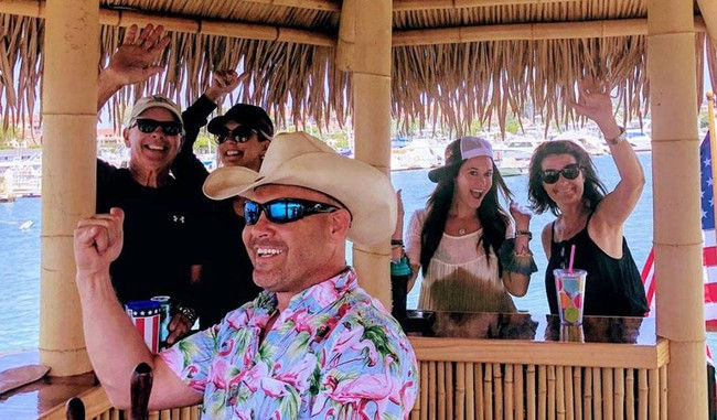 Group of five people enjoying drinks under a bamboo tiki hut at a waterfront marina — man in a cowboy hat and floral shirt in the foreground, friends waving behind the bar with boats and water in the background.
