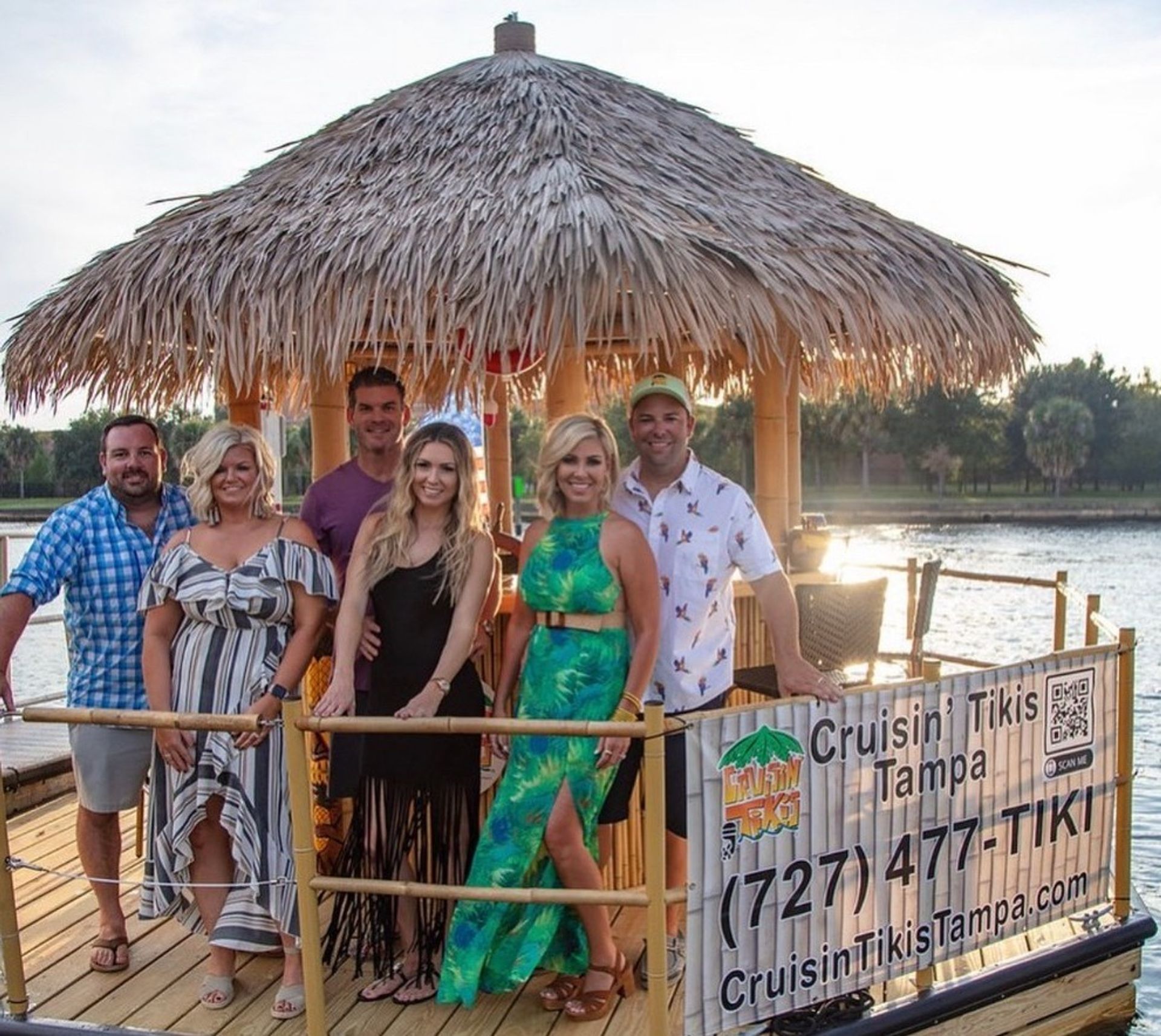 Group of six adults smiling on a tiki-style boat with a palm-thatched roof, docked on a calm waterfront at sunset, wearing casual summer outfits for a tropical-themed outing.