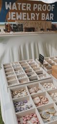 Beach-themed waterproof jewelry bar display at a market: organized compartment trays filled with seashells, pearls, pastel beads and gold-accented shell pendants on a white table.