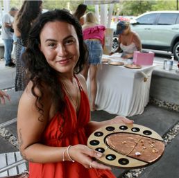 Smiling woman in a red dress holding a round copper jewelry display with rings, earrings and a necklace at an outdoor craft market stall, vendors and shoppers blurred in the background.