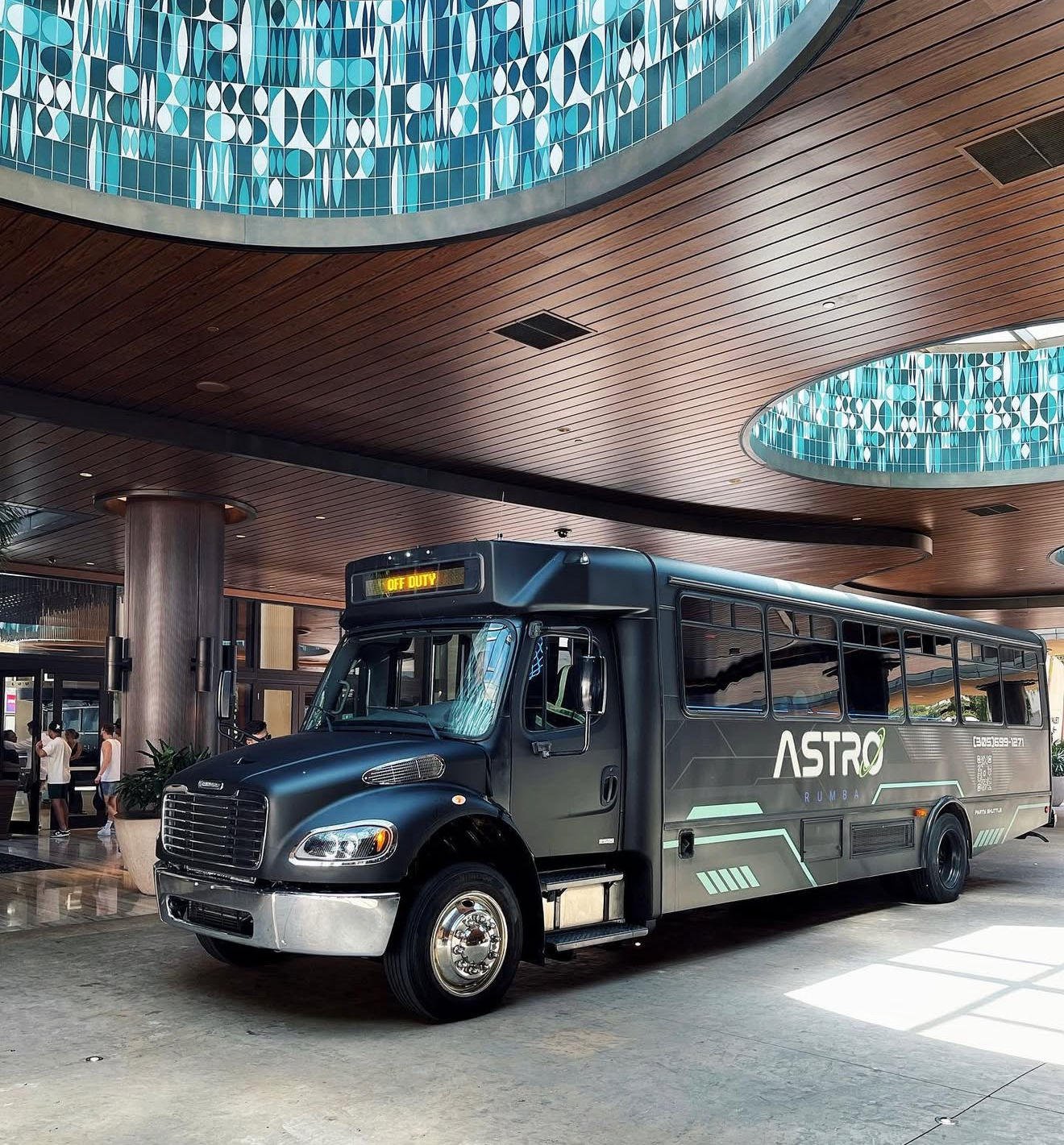 Black shuttle bus parked in a modern hotel lobby/porte-cochere beneath curved wood-paneled ceiling and circular turquoise geometric skylights, digital sign reading "Off Duty".