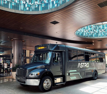 Black shuttle bus parked in a modern hotel lobby/porte-cochere beneath curved wood-paneled ceiling and circular turquoise geometric skylights, digital sign reading "Off Duty".